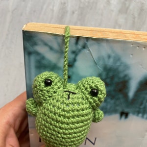May include: A handmade green crochet frog bookmark with black eyes and a small mouth. The frog is attached to a green yarn loop, designed to hang over the top of a book. The bookmark is placed in a book with the title "Siege".
