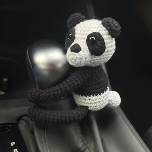 以下が含まれることがあります： 車のシフトレバーに、黒と白の毛糸で編まれたパンダのぬいぐるみが取り付けられています。パンダは黒い目と黒い鼻をしています。