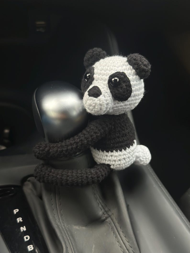 以下が含まれることがあります： 車のシフトレバーに、黒と白の毛糸で編まれたパンダのぬいぐるみが取り付けられています。パンダは黒い目と黒い鼻をしています。