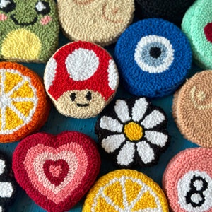 May include: A collection of colorful, round, and heart-shaped crocheted ornaments. The ornaments feature various designs, including a frog, a mushroom, a daisy, a lemon, an evil eye, a pool ball, and a heart.