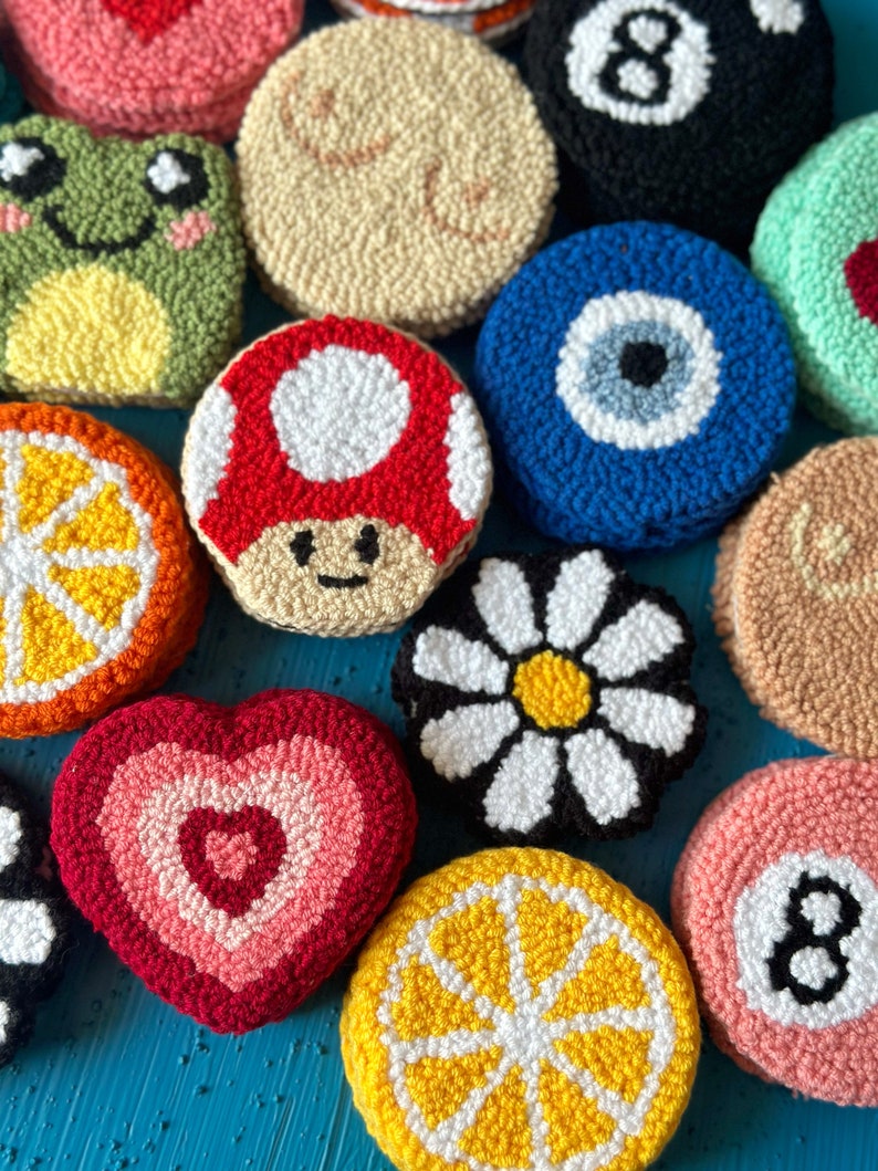 May include: A collection of colorful, round, and heart-shaped crocheted ornaments. The ornaments feature various designs, including a frog, a mushroom, a daisy, a lemon, an evil eye, a pool ball, and a heart.