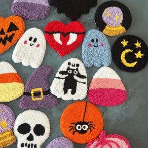 May include: A collection of colorful, textured Halloween-themed coasters. Designs include a bat, ghosts, candy corn, a skull, a witch's hat, a spider, a pumpkin, a heart, and a fortune-telling ball. The coasters are made of a soft, plush material.
