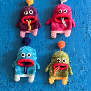 Puede incluir: Cuatro juguetes de peluche de crochet en diferentes colores, cada uno con una gran boca roja, dos ojos negros y una lengua que sobresale. Los juguetes est&aacute;n dispuestos en fila sobre un fondo azul.