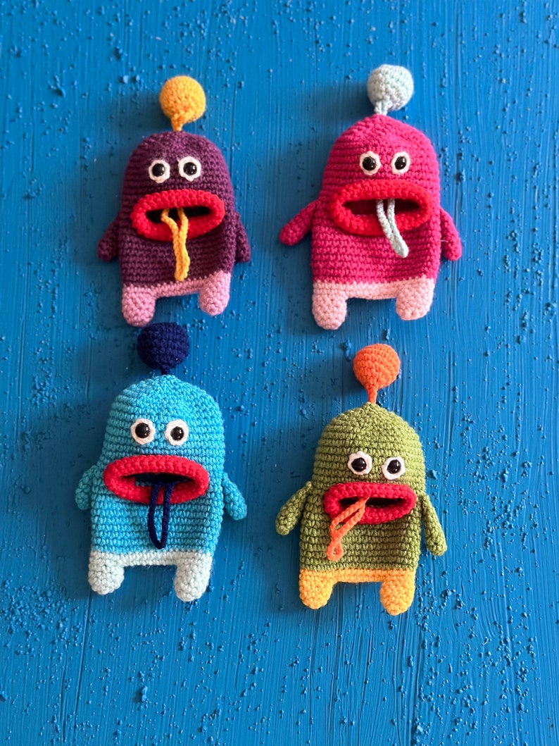 Puede incluir: Cuatro juguetes de peluche de crochet en diferentes colores, cada uno con una gran boca roja, dos ojos negros y una lengua que sobresale. Los juguetes est&aacute;n dispuestos en fila sobre un fondo azul.