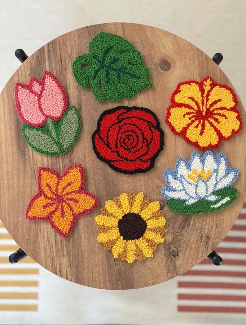 May include: A collection of colorful, embroidered floral coasters. The set includes a pink tulip, a red rose, a yellow hibiscus, an orange star-shaped flower, a yellow sunflower, a blue water lily, and a green leaf. The coasters are arranged on a wooden table.