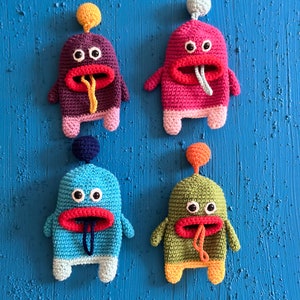 Puede incluir: Cuatro juguetes de peluche de crochet con forma de monstruos con cuerpos y caras de diferentes colores. Los monstruos tienen ojos grandes, bocas rojas y lenguas que sobresalen. Los juguetes est&aacute;n colgados en una pared azul.
