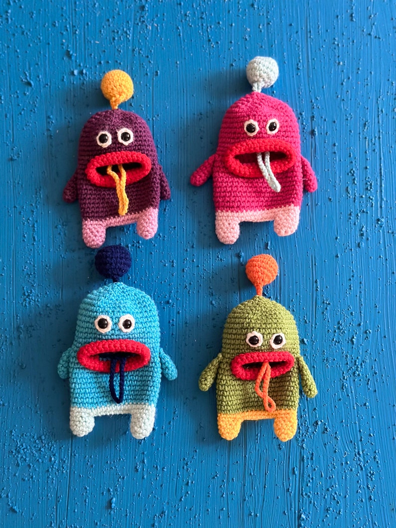 Puede incluir: Cuatro juguetes de peluche de crochet con forma de monstruos con cuerpos y caras de diferentes colores. Los monstruos tienen ojos grandes, bocas rojas y lenguas que sobresalen. Los juguetes est&aacute;n colgados en una pared azul.