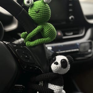 以下が含まれることがあります： 車のステアリングホイールにぶら下がっている緑色の編みぐるみカエルと白黒の編みぐるみパンダ