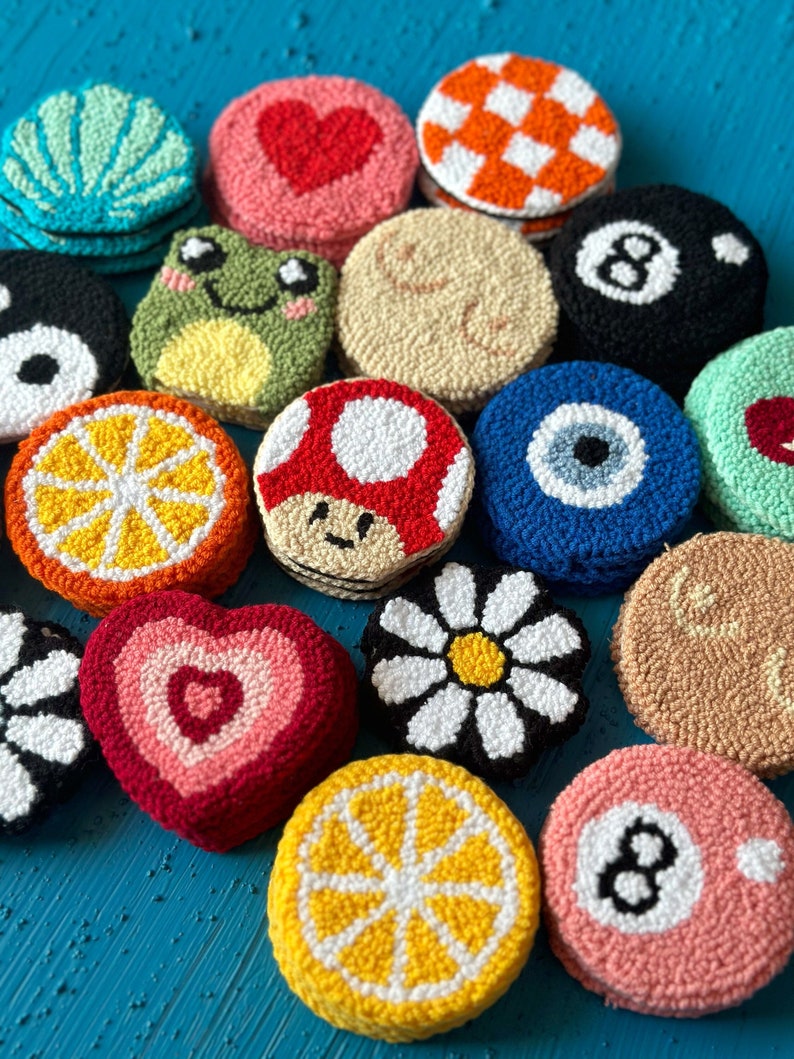 May include: A collection of colorful, round, hand-stitched felt ornaments. The ornaments feature various designs, including a heart, a frog, a mushroom, a daisy, a lemon, an eight ball, and an evil eye.