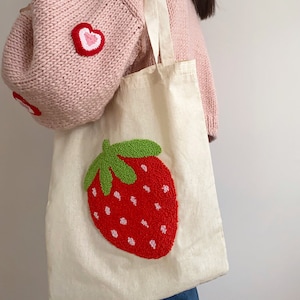 Op de afbeelding: Een witte canvas tote bag met een geborduurd aardbeienontwerp in rood en groen. De tas heeft twee lange hengsels.