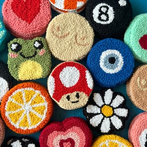 May include: A collection of 14 colorful, round, hand-stitched wool felt ornaments. The ornaments feature various designs, including a heart, a mushroom, a daisy, a lemon, a frog, an eye, a checkered pattern, and the number 8.