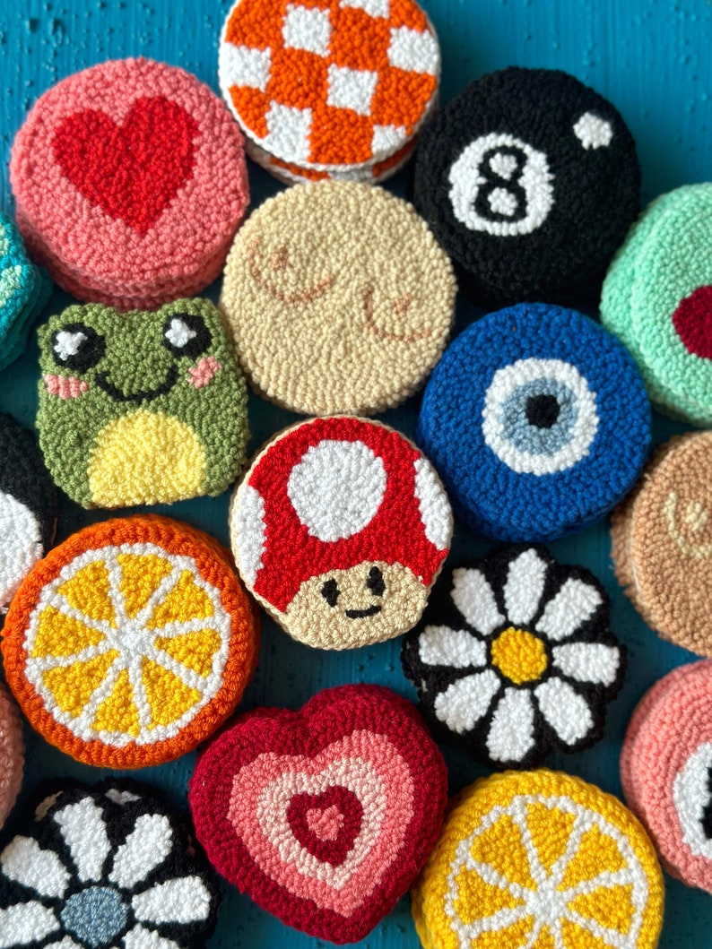 May include: A collection of 14 colorful, round, hand-stitched wool felt ornaments. The ornaments feature various designs, including a heart, a mushroom, a daisy, a lemon, a frog, an eye, a checkered pattern, and the number 8.