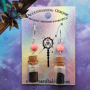 May include: A pair of earrings featuring small glass bottles filled with black strips of paper, each bottle topped with a cork and a pink stone charm. The earrings are displayed on a card with the text "Accentuating Unique Because Ordinary is for NPCs".