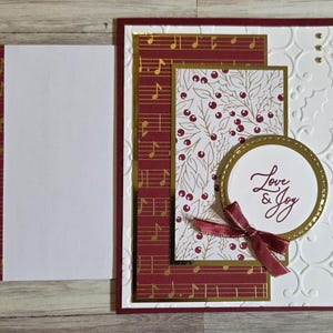 May include: A handmade greeting card featuring a white embossed background with holly patterns, a maroon panel with gold musical notes, a floral print with red berries, and a circular center piece with the text Love & Joy and a maroon ribbon.