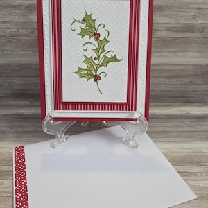 May include: A rectangular greeting card featuring a central white panel with a green holly and red berry illustration, layered over a red and white striped background and a white embossed border, accompanied by a matching white envelope.