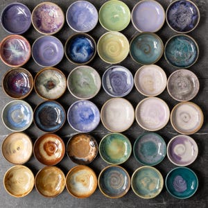 May include: A collection of thirty-six small, round ceramic bowls arranged in a grid. Each bowl features a unique reactive glaze finish in various colors including shades of blue, purple, green, yellow, brown, and white, with swirling patterns.