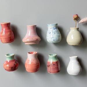 May include: Eight small ceramic vases arranged in two rows. The vases feature various finishes including solid pink, speckled blue, cream, and multi-colored glazes. One cream-colored vase contains a small dried pink flower and a feathery plume.