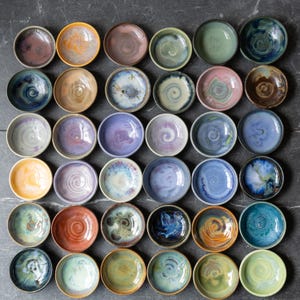 Op de afbeelding: Een verzameling kleine, handgemaakte keramische kommen in verschillende kleuren, waaronder blauw, groen, oranje en paars. Elke kom heeft een uniek glazuurpatroon en een enigszins onregelmatige vorm, wat suggereert dat ze handgemaakt zijn. De kommen zijn in een rasterpatroon gerangschikt.