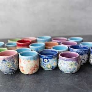 May include: A selection of small, hand-painted ceramic cups. The cups display floral patterns in various colours, including blue, pink, and green. The cups are arranged on a dark surface.