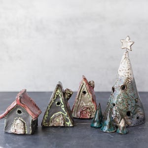 May include: A collection of ceramic houses and trees in various shades of green, brown, and red. The houses have small windows and doors, and the trees have a star on top. The houses and trees are arranged on a dark gray surface.