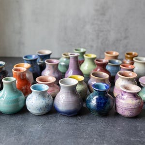 May include: A selection of small, handmade ceramic vases in a range of colours, such as green, blue, orange, and purple. Each vase has a unique form and glaze, creating a diverse and artistic display. The vases are arranged on a dark surface.