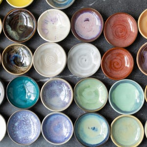 May include: Overhead shot of a collection of small, handmade ceramic bowls in various colors. The bowls have a glossy finish and are arranged in a grid pattern. Colors include teal, purple, green, brown, and white.