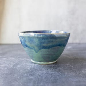 May include: A ceramic bowl featuring a smooth, rounded shape with a glossy glaze. The exterior displays a blended color palette of deep navy blue at the top, transitioning into a vibrant teal and earthy green toward the base, with visible drips.