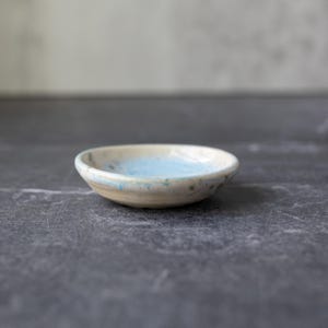 May include: A small, handmade ceramic bowl with a light blue interior and a beige exterior. The bowl has a shallow, rounded shape and is likely used for holding small items. The glaze has a speckled appearance, adding to its unique, handcrafted aesthetic.