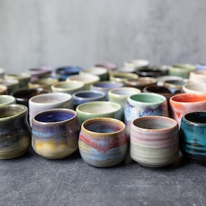 May include: A large collection of small ceramic cups featuring a variety of reactive glazes in colours including blue, green, pink, orange, and purple. Each cup has a rounded, handleless shape with a smooth finish and visible colour gradients.