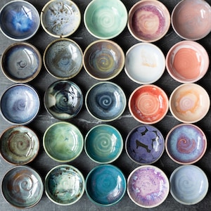 May include: A collection of twenty-five small, round ceramic bowls arranged in a five-by-five grid. Each bowl features a unique glaze, including shades of blue, green, pink, purple, and orange, with various speckled, swirled, and solid finishes.
