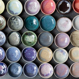 Peut inclure: Une collection de trente petits bols en céramique ronds disposés en grille. Chaque bol présente une finition émaillée unique dans des tons variés de bleu, violet, rose, vert, jaune et marron, avec des motifs en spirale visibles.