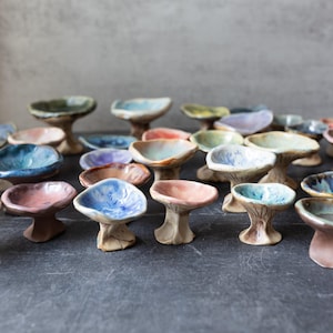 May include: A collection of small, handmade ceramic bowls with a pedestal base. The bowls are in various colors, including blue, green, pink, and yellow. The ceramic bowls are arranged on a dark surface.