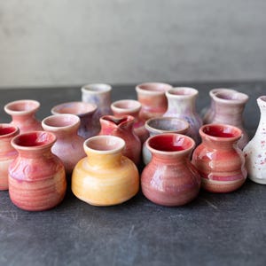 May include: A collection of small, handmade ceramic vases in various shapes and sizes. The vases feature a range of colors, including red, pink, yellow, and white, with textured and glazed finishes. Some have a speckled appearance, while others have smooth surfaces.