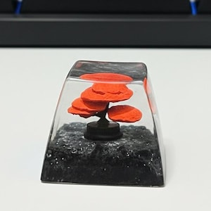 May include: A clear resin keycap with a miniature orange tree design. The tree has a black trunk and sits on a bed of black, glittery material. The keycap is rectangular and sits in front of a keyboard.