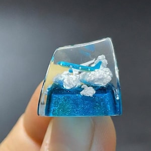 May include: A clear, rectangular resin keycap featuring a miniature blue airplane flying above fluffy white clouds. The base of the keycap is a vibrant blue, resembling water. The keycap is held by a person's fingers.