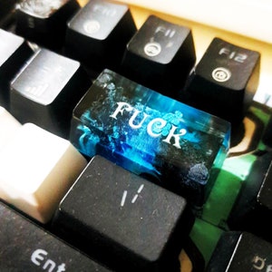 Puede incluir: Una tecla de teclado de resina azul con la palabra "FUCK" impresa en letras blancas.