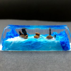 May include: A miniature scene of two small wooden ships sailing on blue water with a small treasure chest in the middle. The scene is encased in clear resin.