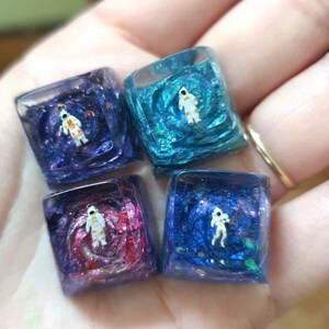 May include: Four square resin keycaps with miniature astronaut figures suspended within swirling, colorful space-themed designs. The keycaps feature shades of blue, purple, and pink, with glitter accents. Each keycap has a different space scene.