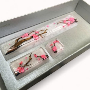 May include: A set of three clear resin keycaps featuring a delicate cherry blossom design with pink flowers and brown branches. The keycaps are displayed in a white box.