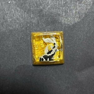 May include: A square, gold-colored decorative object with a koi fish design. The koi fish is black, white, and silver, and appears to be swimming in a golden liquid. The object has a textured surface and is likely a decorative item.