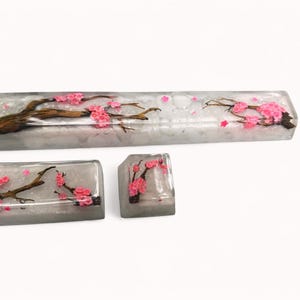 Peut inclure: Un ensemble de trois touches de clavier avec un fond blanc et un motif d&eacute;licat de fleurs de cerisier roses. Les touches sont en r&eacute;sine transparente et ont une finition scintillante.