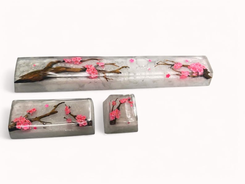 Peut inclure: Un ensemble de trois touches de clavier avec un fond blanc et un motif d&eacute;licat de fleurs de cerisier roses. Les touches sont en r&eacute;sine transparente et ont une finition scintillante.