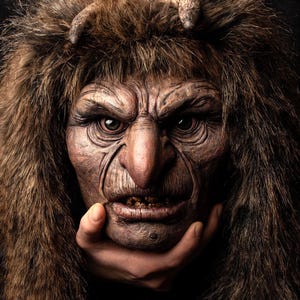 May include: A detailed mask of a grotesque, aged face with a large nose, deep wrinkles, and a fur headdress. The mask is held by a hand, and the overall color palette is brown and dark.