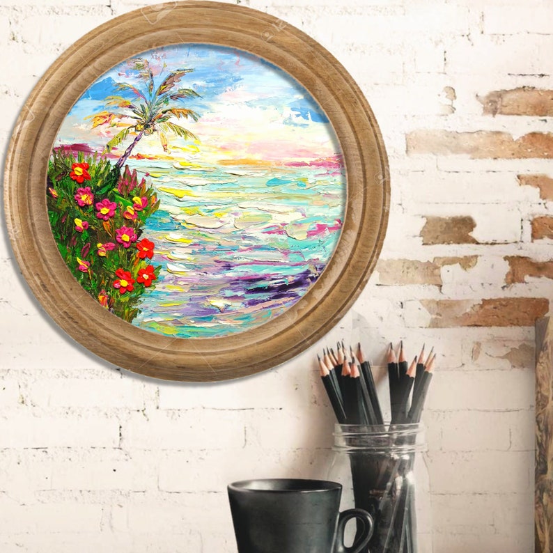 Puede incluir: Una pintura redonda enmarcada que representa una escena de playa tropical con una palmera, flores rosas y rojas y agua azul. La pintura est&aacute; en una pared de ladrillo blanco.