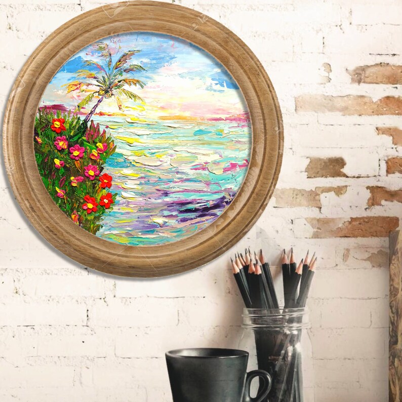 Puede incluir: Una pintura redonda enmarcada que representa una escena de playa tropical con una palmera, flores rosas y rojas y agua azul. La pintura est&aacute; en una pared de ladrillo blanco.
