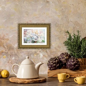 May include: A still life featuring a framed painting with a gold frame, a white teapot, lemons, artichokes, and rosemary. The painting has a floral theme. The scene is set on a wooden surface.