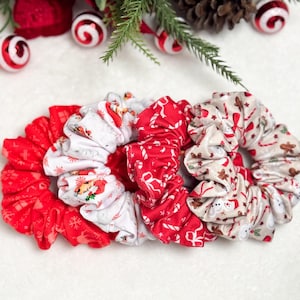 May include: A collection of festive scrunchies in red, white, and beige. The scrunchies feature holiday-themed patterns, including candy canes, gingerbread men, and reindeer. These hair accessories are perfect for adding a touch of holiday cheer to any hairstyle.