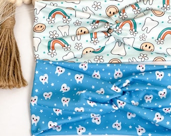 May include: Two fabric headbands with a tooth pattern. One headband is blue with white teeth and stars. The other headband is white with blue rainbows, flowers, and smiley faces.