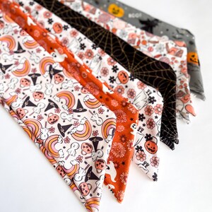 Może przedstawiać: Kolekcja materiałowych opasek do włos&oacute;w o tematyce Halloween. Wzory obejmują dynie, duchy, tęcze, pajęczyny i nadruki kwiatowe. Kolory to pomarańczowy, czarny, biały i szary. Opaski są ułożone w kształt wachlarza.