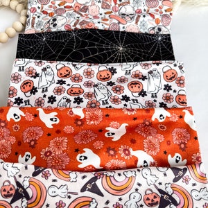 Może przedstawiać: Stos materiałowych opasek do włos&oacute;w z motywem Halloween. Opaski mają r&oacute;żne wzory, w tym duchy, dynie, pajęczyny i kwiatowe wzory. Kolory to czarny, biały, pomarańczowy i szary. Opaski prawdopodobnie na uroczystą okazję.